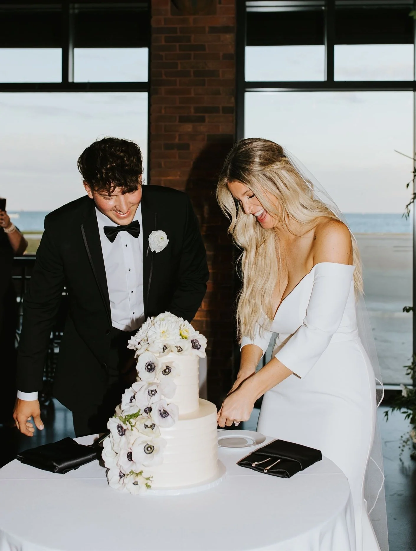Bride and groom smiling as bride cuts into wedding cake made by ECBG Studio with fresh white flowers cascading from top tier