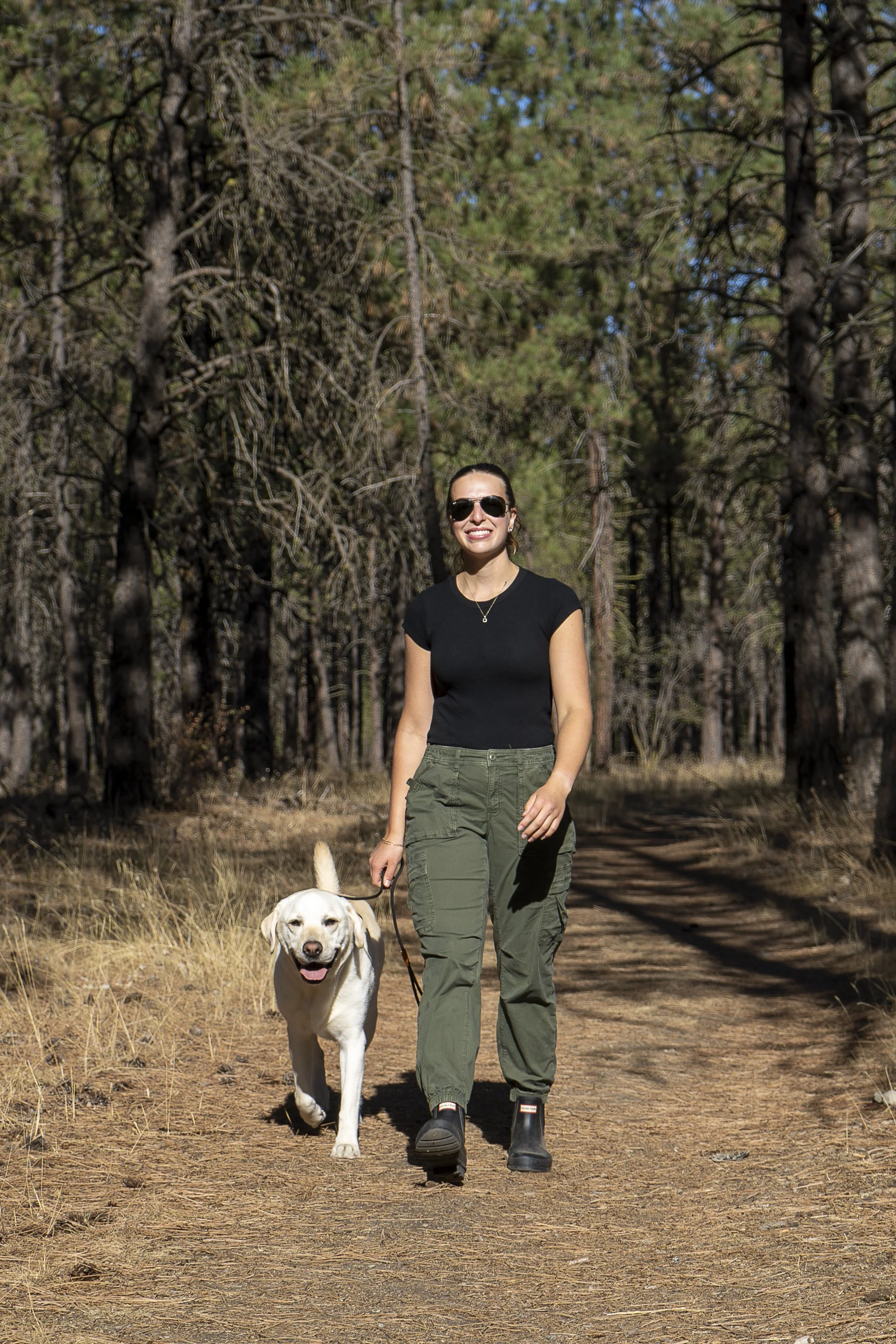 A woman smiling and wearing sunglasses, a black t-shirt, and green cargo pants, walking a dog on a trail through a forest.