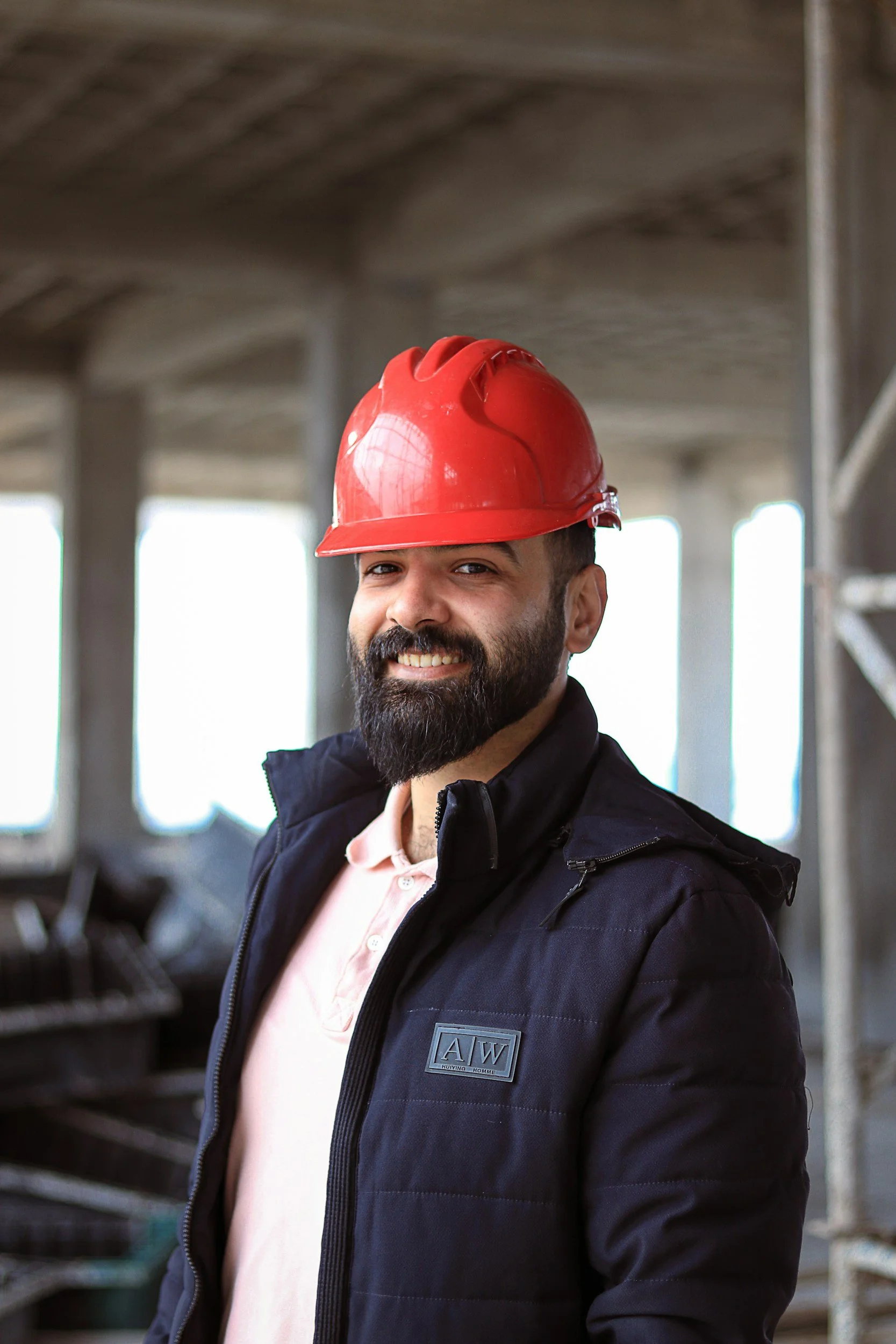 A man with a beard and mustache wearing a red safety helmet and a black jacket, standing on a construction site with scaffolding and blurred background.