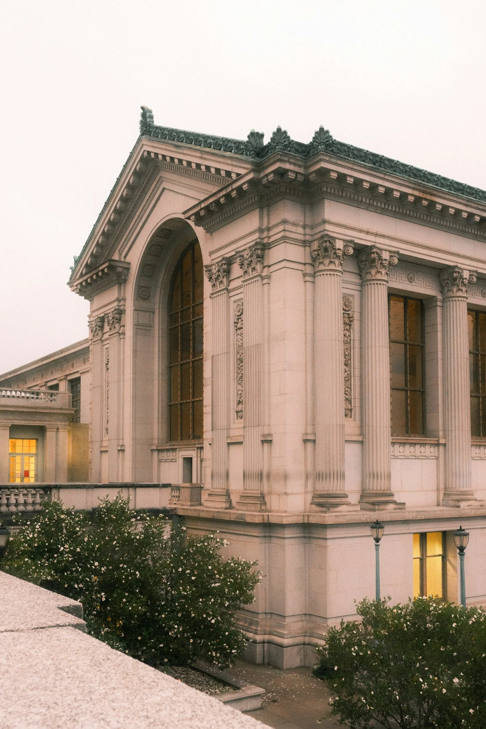 The image shows a classical-style building with ornate architectural details, large windows, and green roof decor, likely a historic or government structure, with shrubbery and a lamp post visible in front.