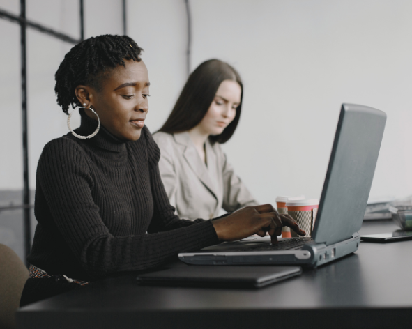 Two women work at a desk with a laptop in an office setting.