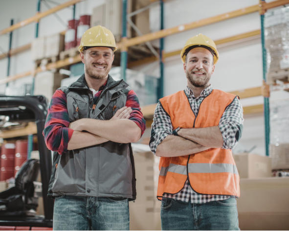 Two male workers wearing yellow safety helmets and casual work clothes standing inside a warehouse with shelves and boxes in the background.