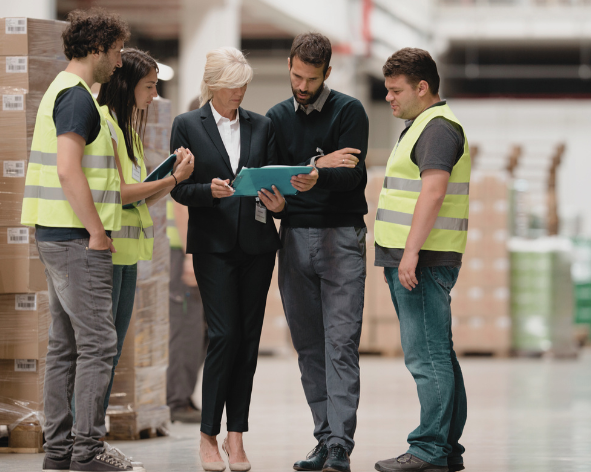 Group of five people, two men and three women, standing in a warehouse, viewing a clipboard, with pallets and boxes in the background.