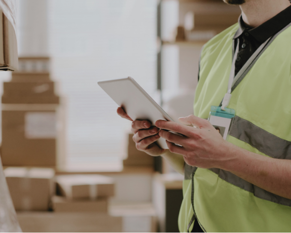 Person in high-visibility vest holding a tablet in a warehouse with cardboard boxes.