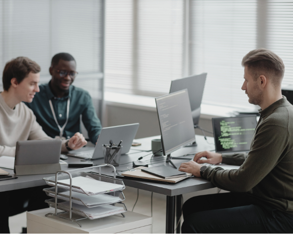 Three people working in a modern office with multiple computer monitors, one person coding while two others observe and discuss.