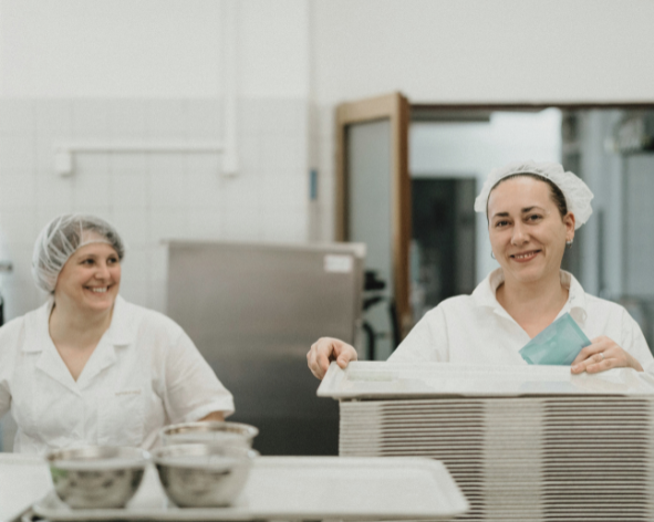 Two women in a commercial kitchen, wearing white uniforms and hairnets, smiling as they prepare food.