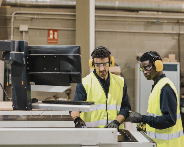 Two workers in yellow safety vests, ear protection, and gloves operating a large piece of machinery in an industrial warehouse.