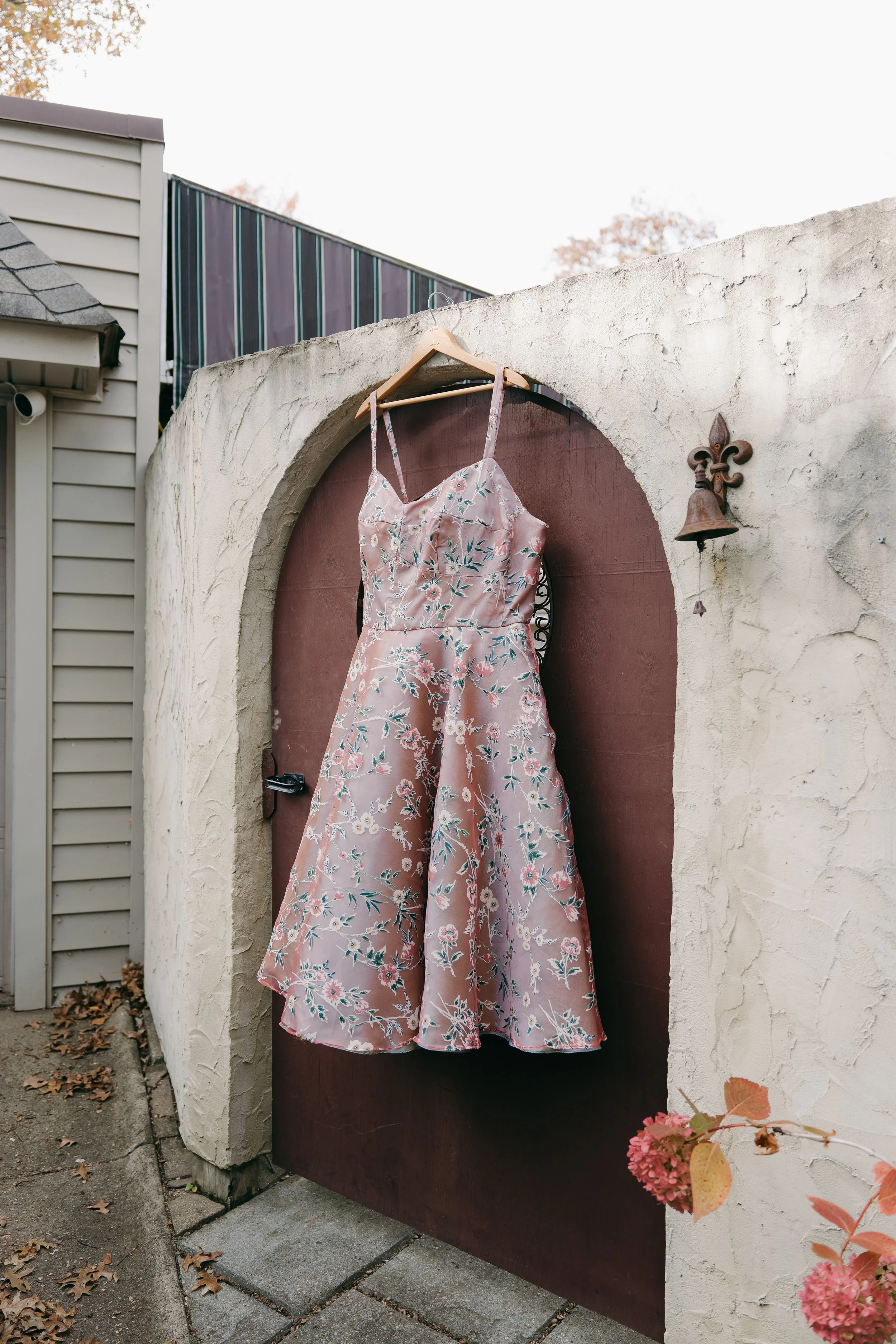 A pink floral dress hangs on a wooden hanger on a door within an arched alcove on a white stucco wall. A small bell with a fleur-de-lis decoration is mounted on the wall to the right of the door, and pink flowers are visible in the lower right corner.