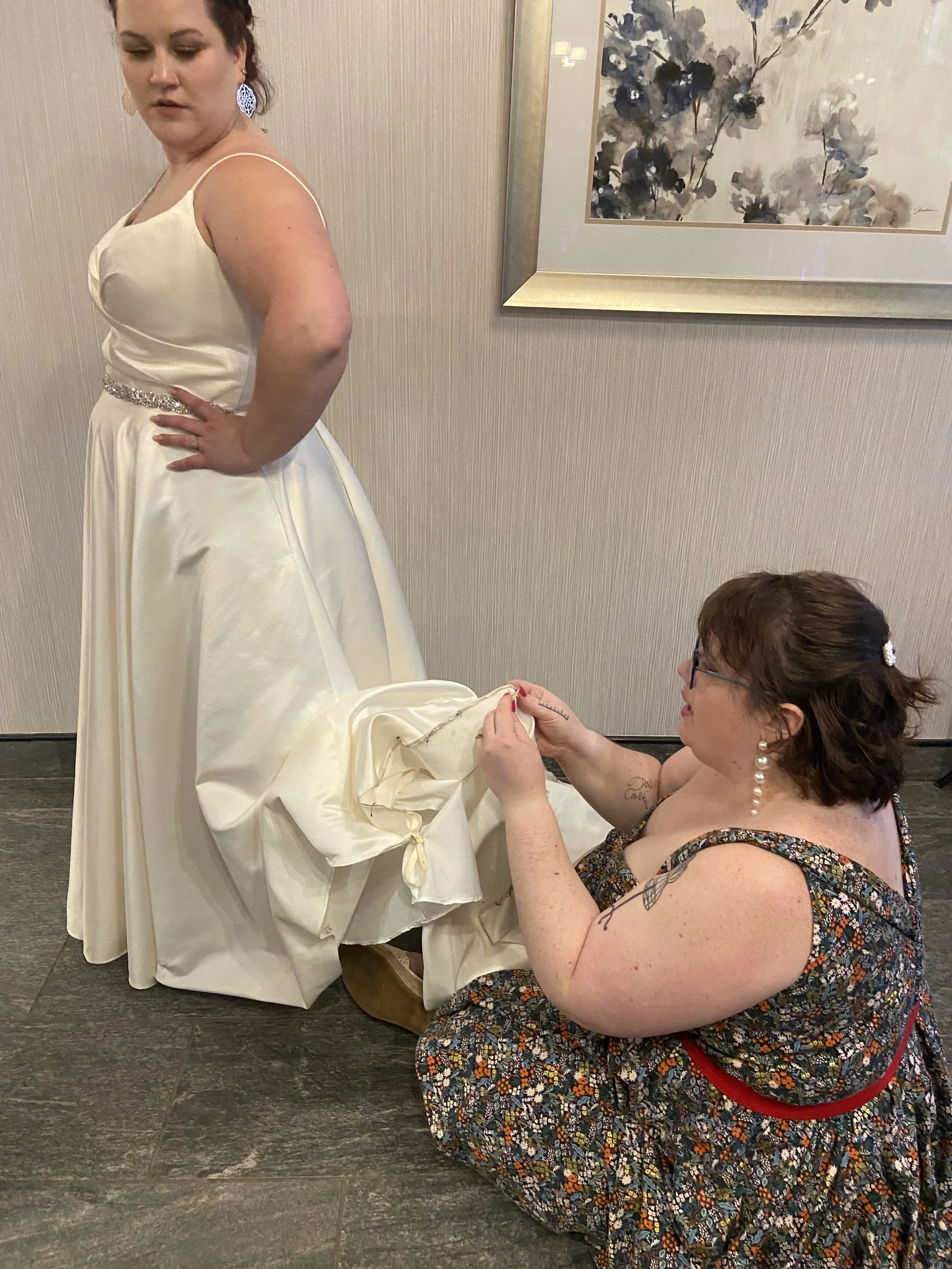 A woman in a wedding dress is having her dress adjusted by another woman crouching on the floor.