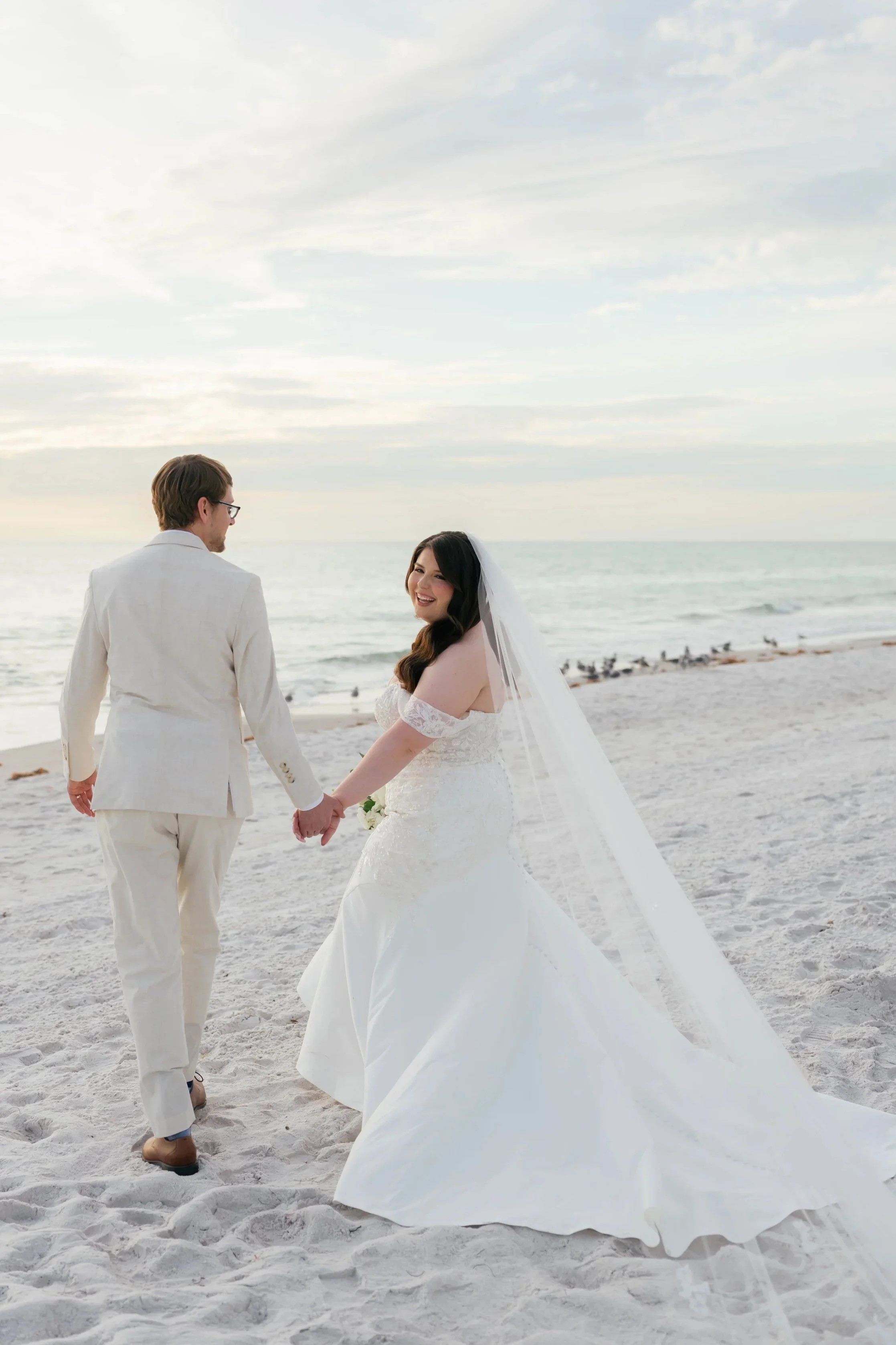 A bride and groom holding hands on the beach during sunset, with the ocean in the background.