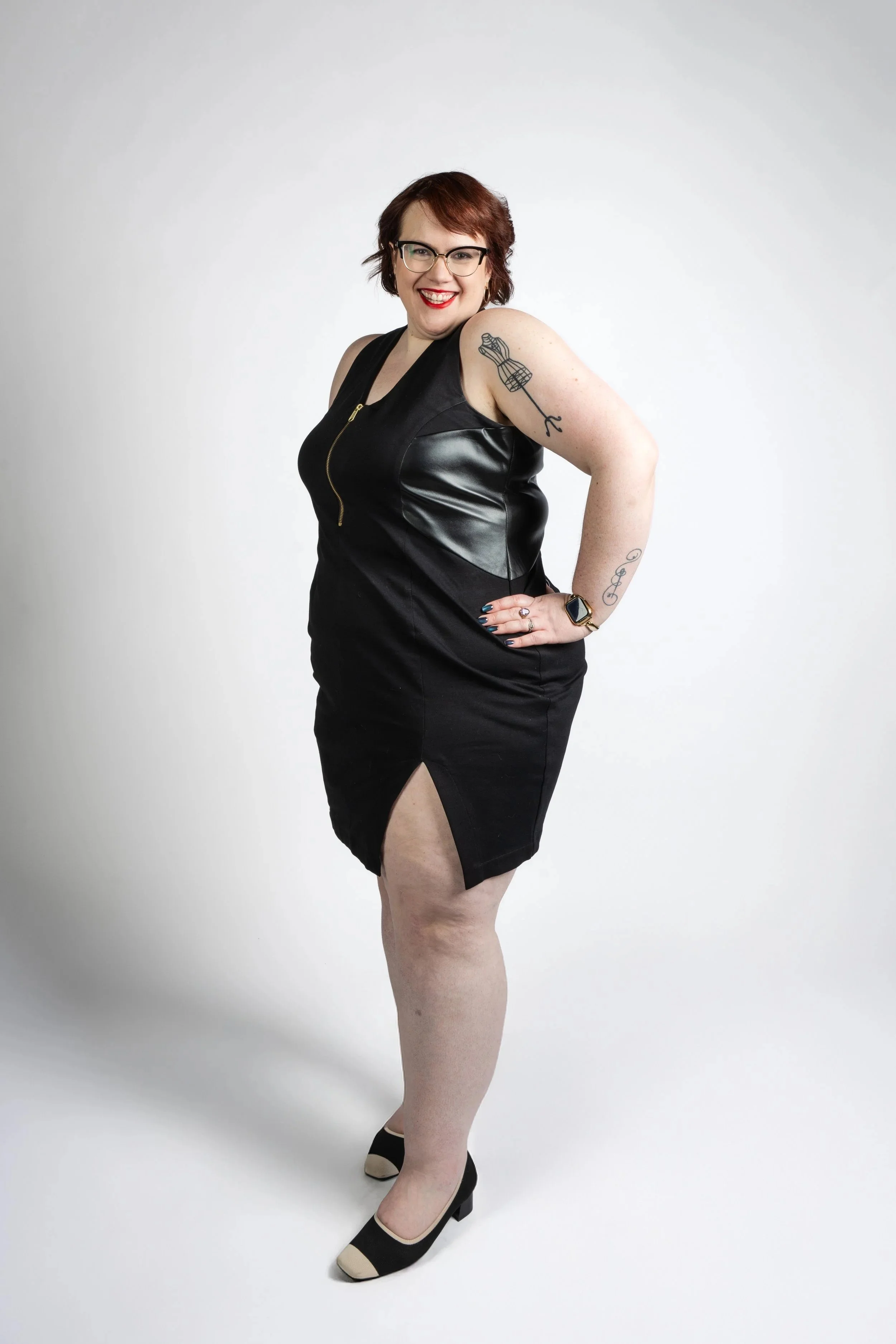 A woman with short red hair and glasses wearing a black dress with leather accents, standing against a plain white background, smiling with her hand on her hip.