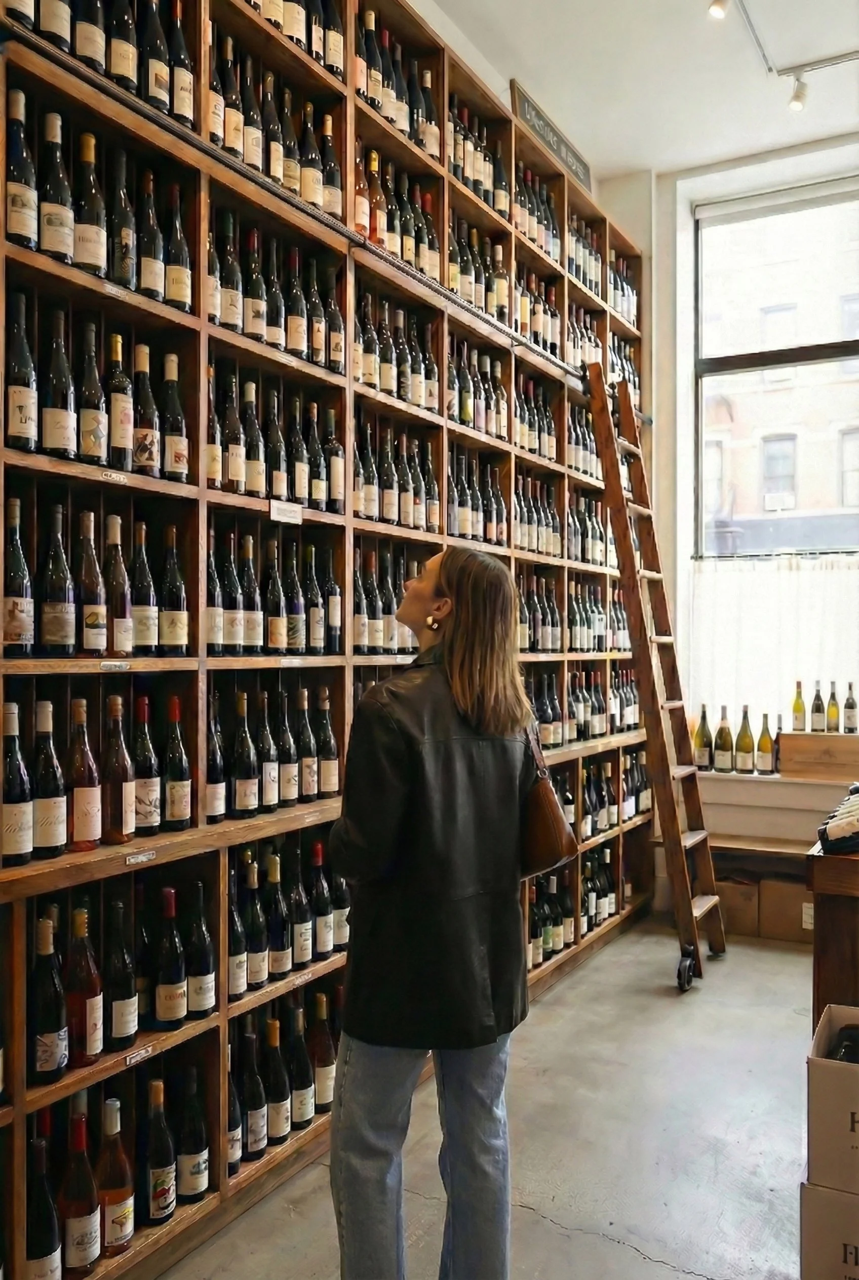 A woman stands in a wine shop, looking at shelves filled with bottles of wine. There is a ladder leaning against the shelves near a large window, with sunlight coming through the window.