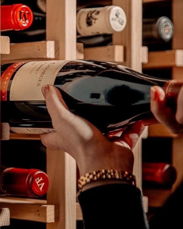 A person holding a bottle of red wine in a wine cellar or wine store with wooden wine racks filled with wine bottles.