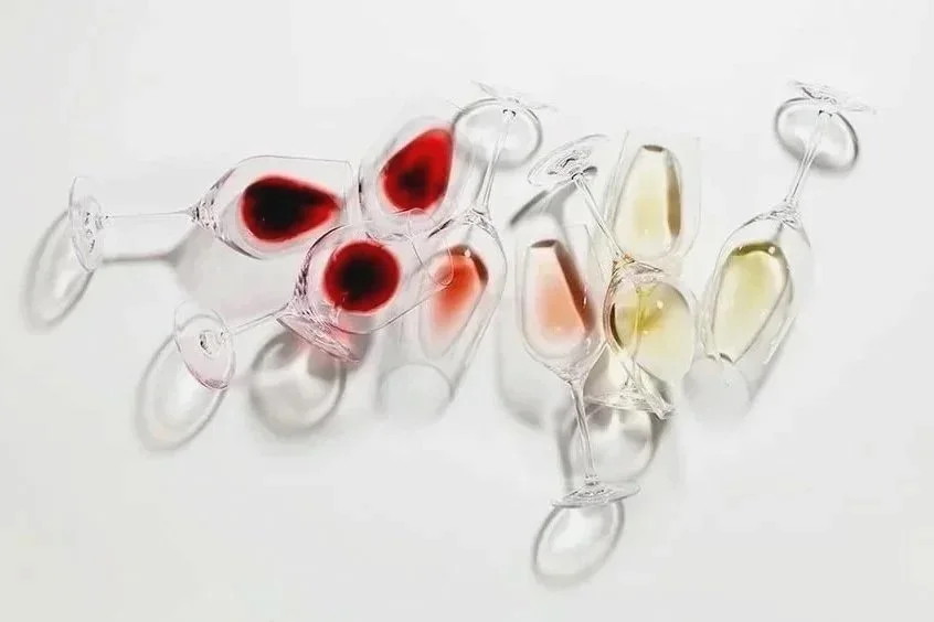 Several empty champagne flutes lying on a white surface, some filled with red, pink, and white wine.