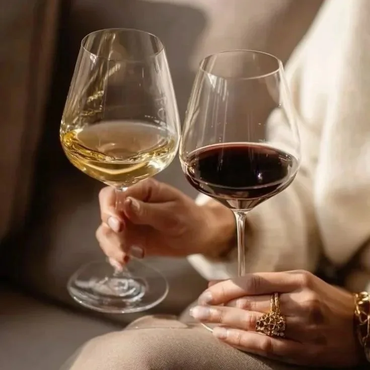 Two glasses of wine, one white and one red, held by a person with jewelry, in a cozy indoor setting.