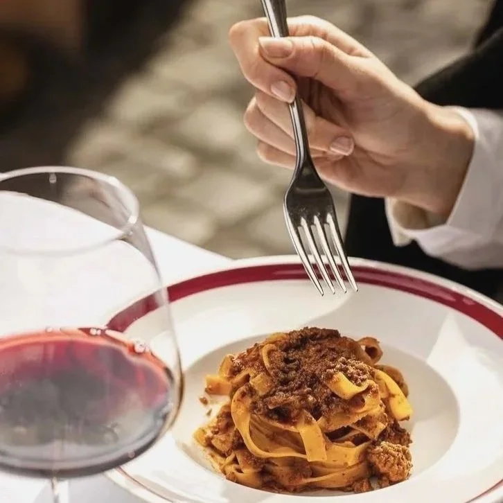 Person holding a fork above a plate of spaghetti with meat sauce, with a glass of red wine on a white tablecloth.
