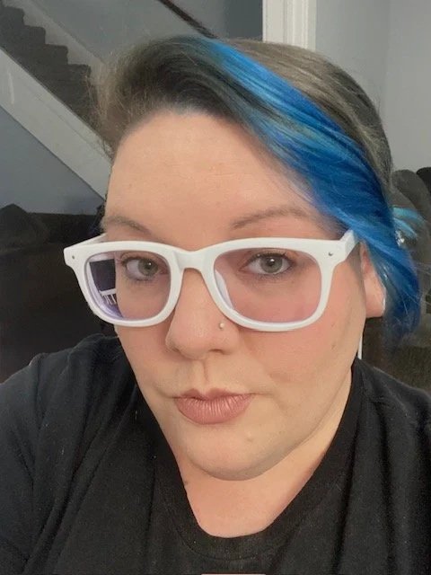 A person with short hair, styled with a blue streak, wearing white glasses, a nose piercing, and a black top, sitting indoors.