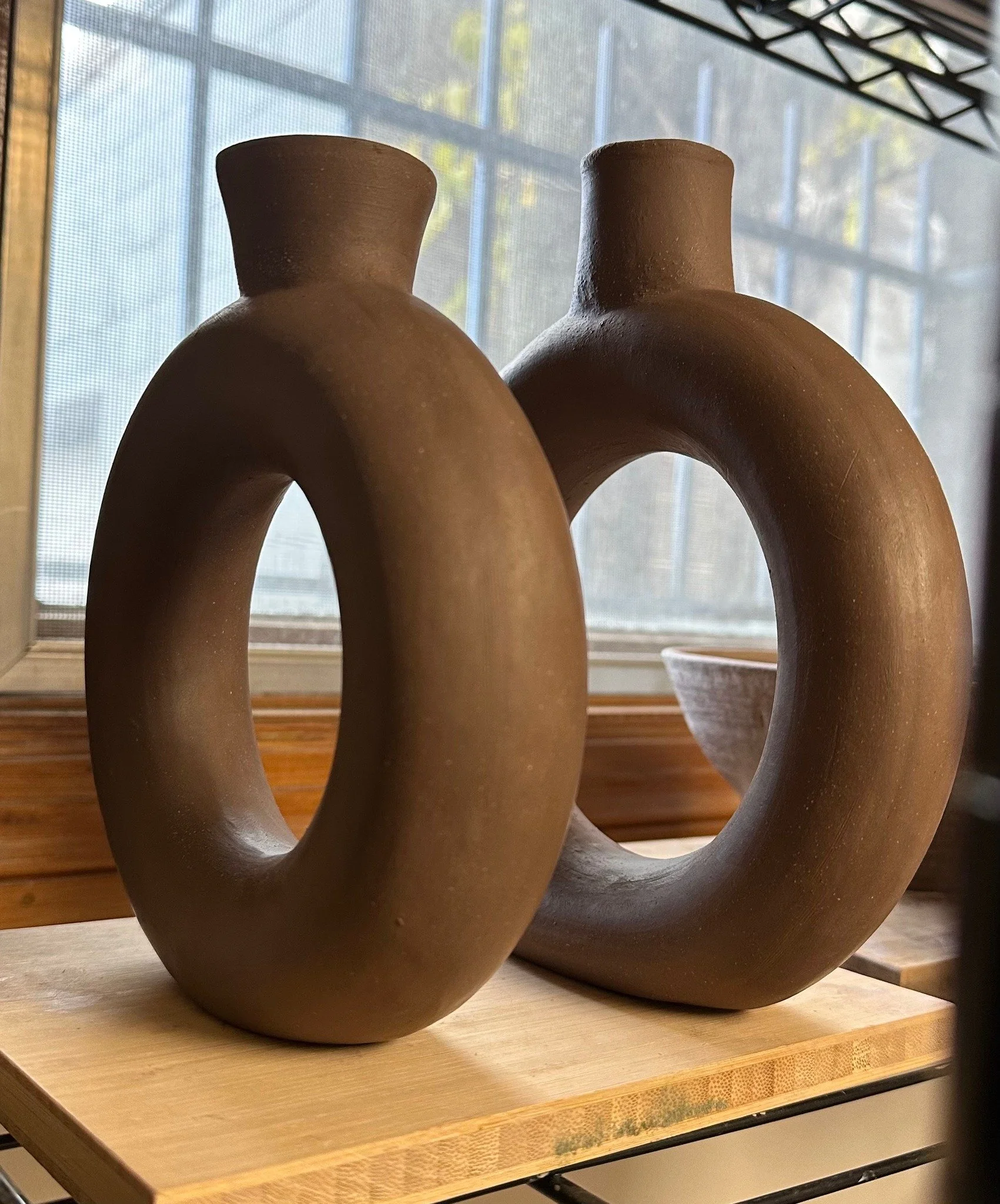 Two ceramic vases with circular openings and narrow necks, placed on a wooden shelf near a window.