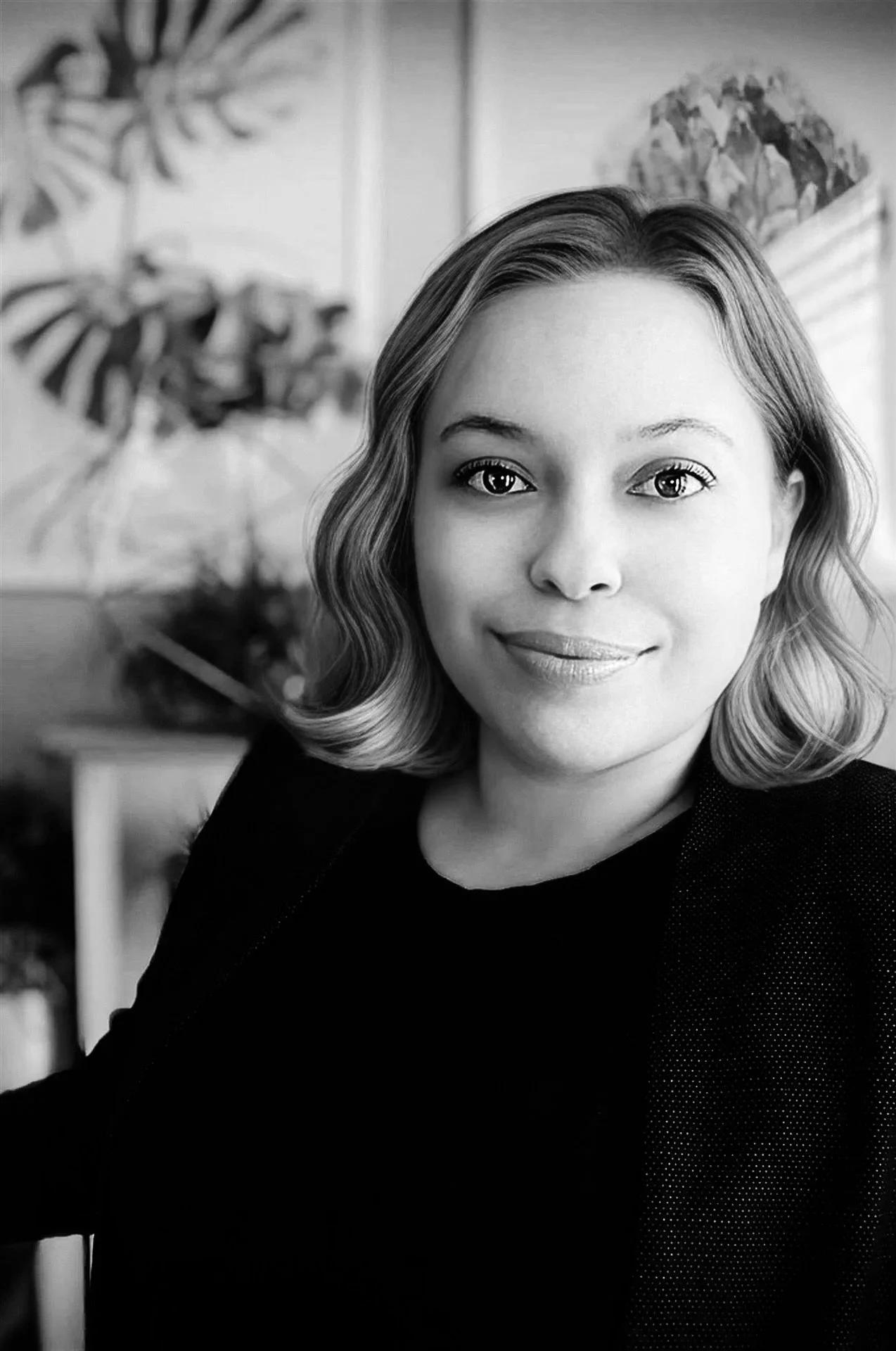 Black and white portrait of Giselle Herrera, the founder of Impact Email Design, a woman with shoulder-length wavy hair, smiling softly, wearing a dark blazer, in an indoor setting with plants and artwork in the background.