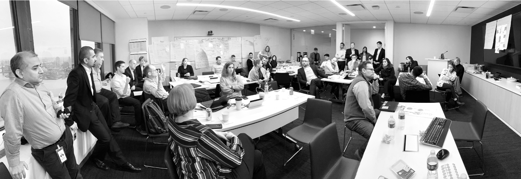A large conference room filled with people attending a meeting or presentation. Some are seated at tables with laptops and notebooks, while others are standing near the back or near the walls. There is a large window on one side of the room showing a city skyline. The front of the room has a podium and a large screen with some papers and notes posted on the wall.