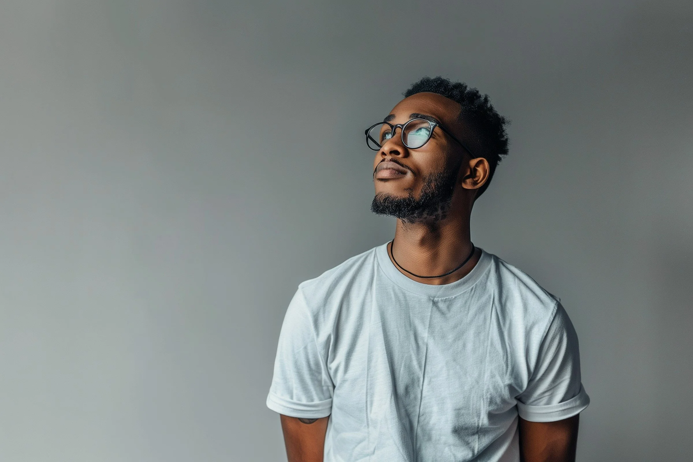 A young African American man with glasses, a beard, and short curly hair, wearing a white T-shirt and a black choker, looking upwards with a thoughtful expression against a plain light gray background.