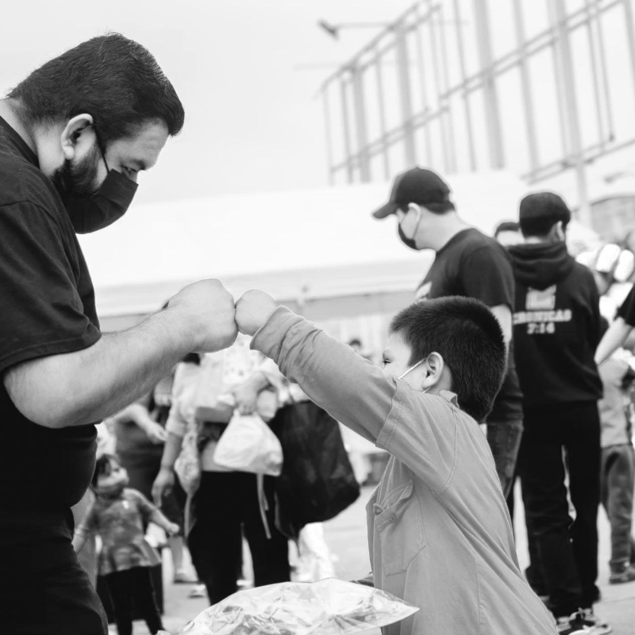 A man and a young boy, both wearing masks, giving each other a fist bump outdoors at a charity event.