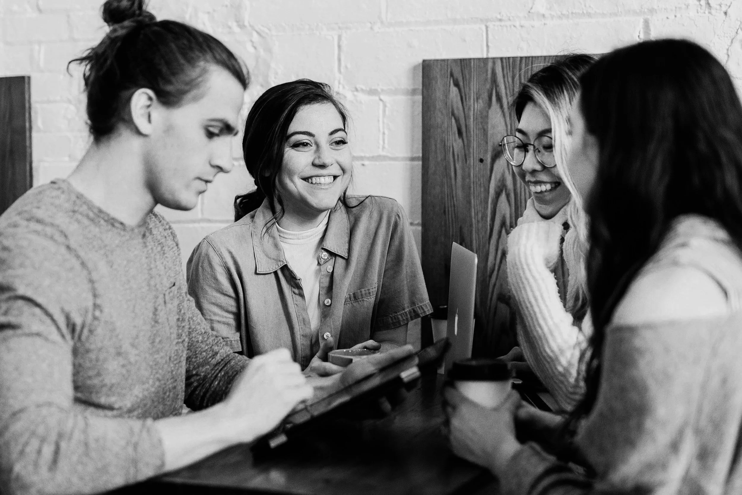 Four women sitting around a table, looking at a laptop and talking, with one woman holding a tablet, in a casual setting.