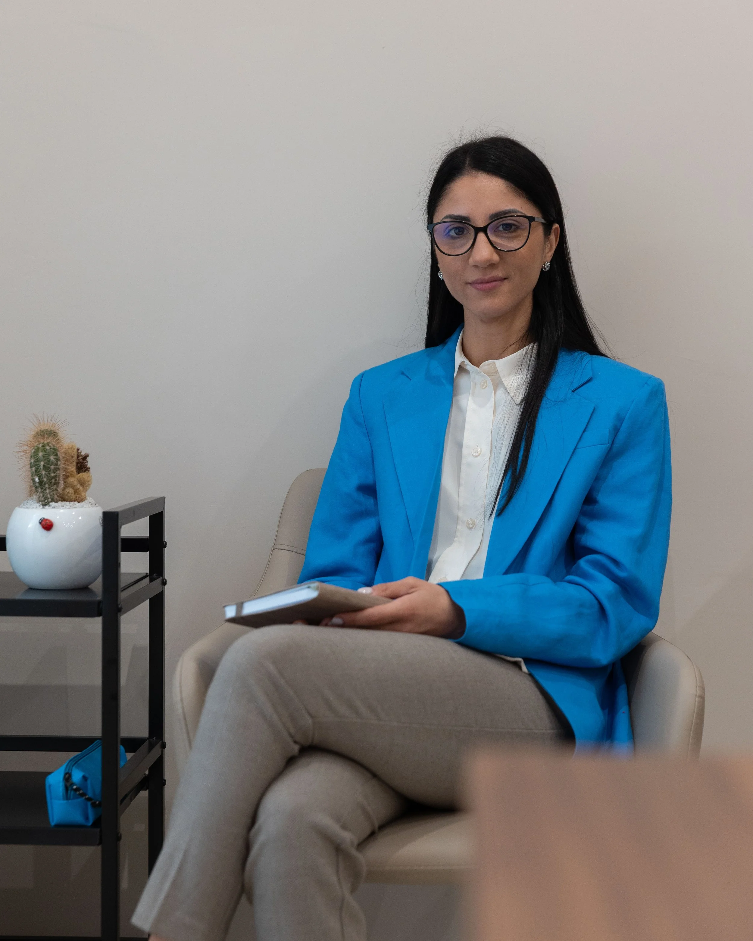 Una donna seduta su una poltrona, vestita con una giacca blu e un camicetta bianca, con occhiali, in un ambiente d'ufficio, che tiene in mano un bloc notes.