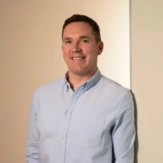 A smiling man in a light blue button-up shirt standing against a beige and white wall.