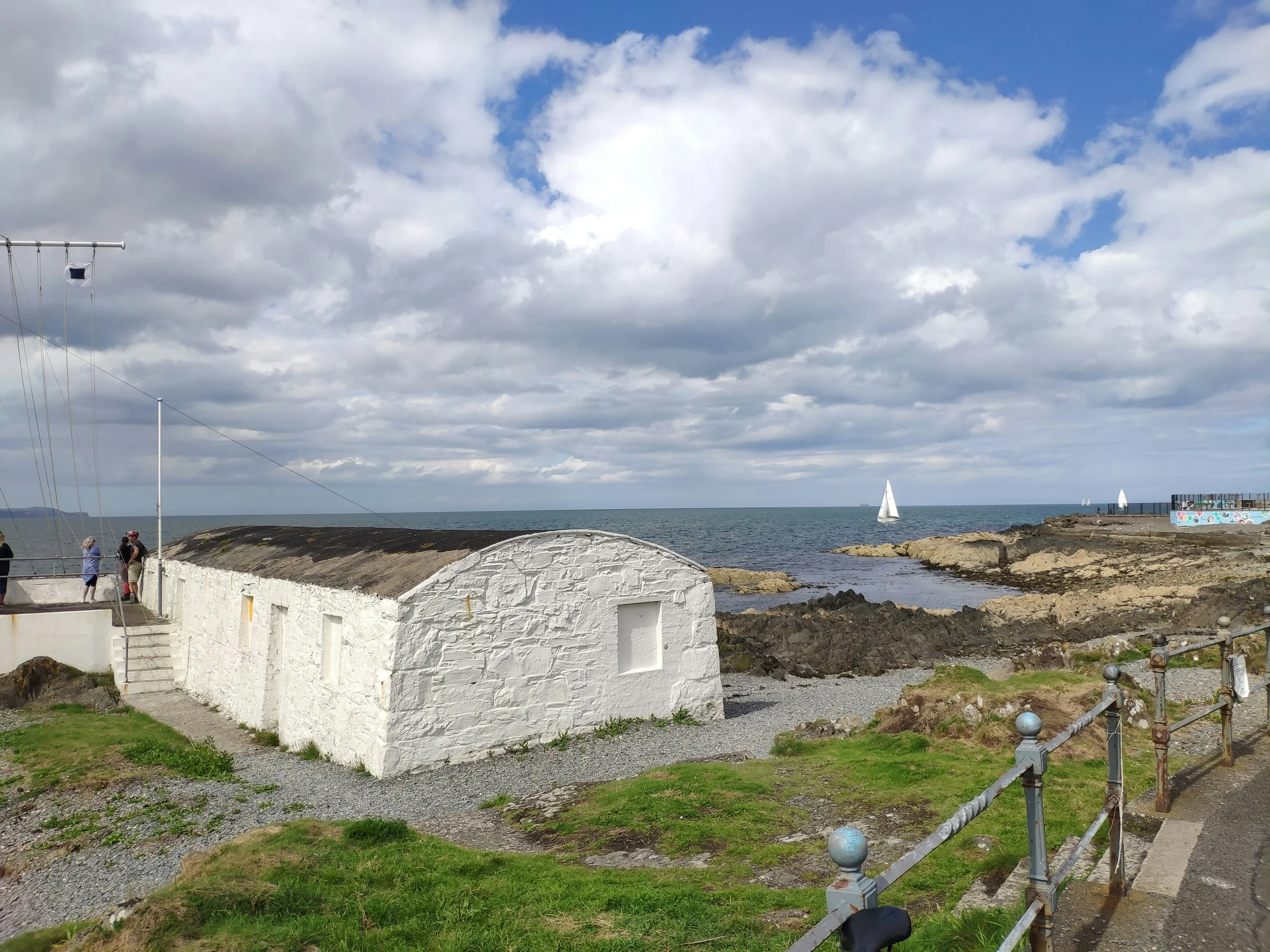 A seaside scene with a white stone building on a grassy area, rocky shoreline, and sailboats on the water under a partly cloudy sky.