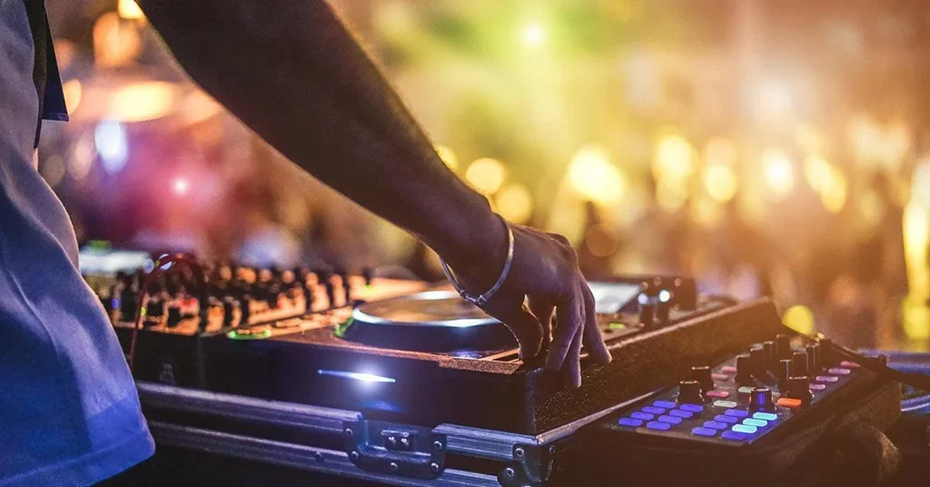 A DJ is playing music at an event, with a focused hand adjusting equipment on a DJ mixer surrounded by various controls and colorful lighting effects.