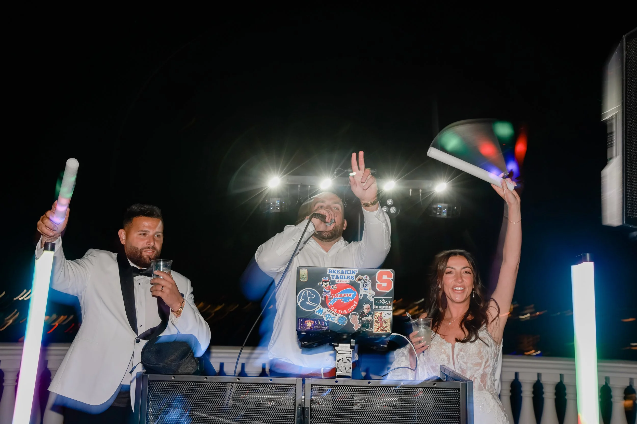 Three people at a party, with two men in tuxedos and a woman in a white dress, partying with glow sticks and a DJ at the center during nighttime.