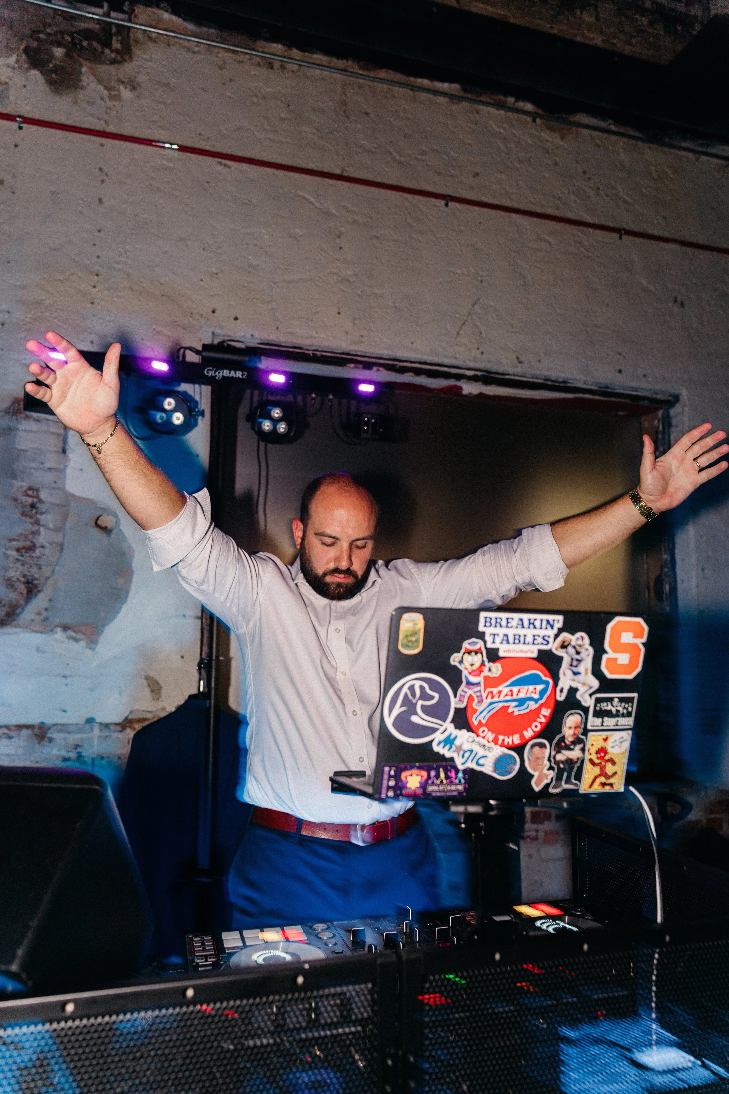 A DJ with a beard and short hair wearing a white shirt with rolled-up sleeves, standing at a DJ booth with arms raised, in a dimly lit venue with brick wall and colorful stickers on the laptop.