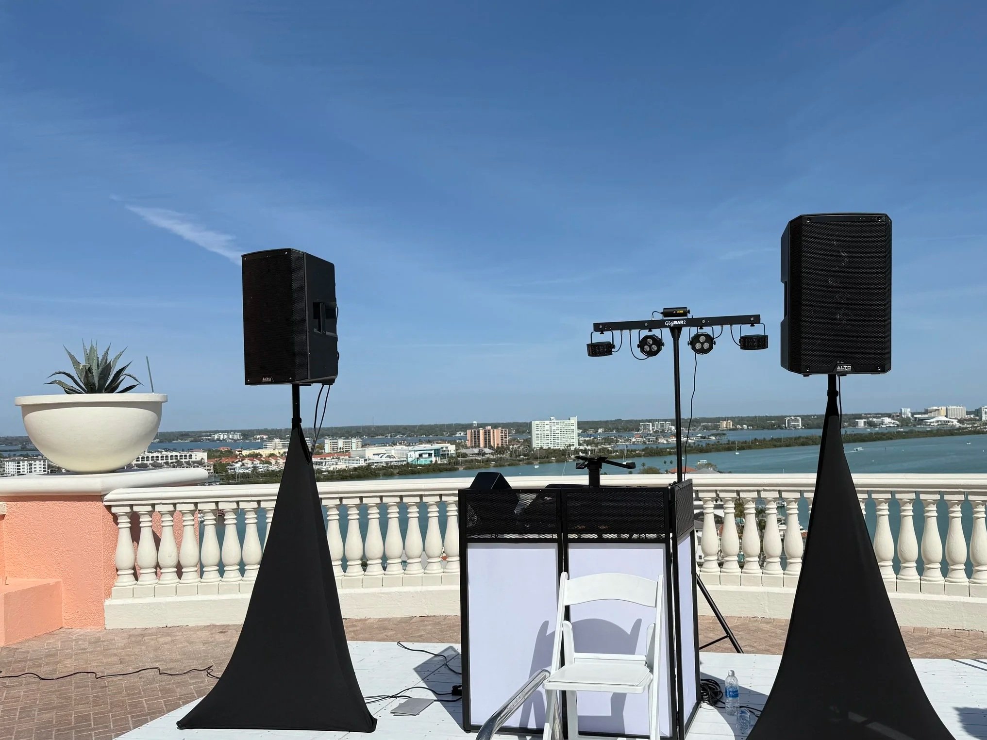 Outdoor stage setup with two large speakers, a DJ console, lighting equipment on a stand, a white chair, and a potted plant, overlooking a city skyline and water body under a blue sky.