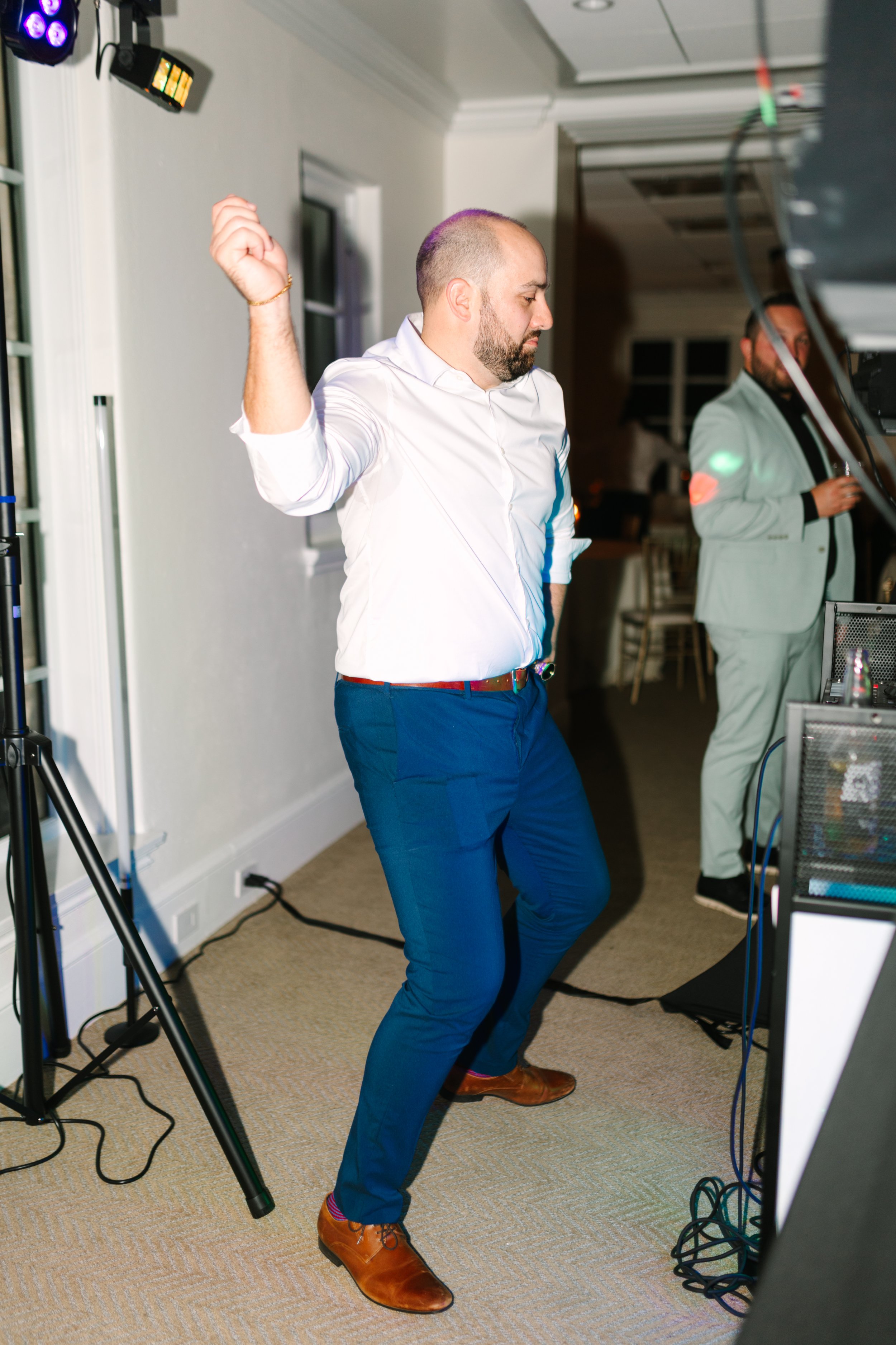 A man in a white shirt and blue pants dancing at an indoor event, with a man in a light suit in the background near DJ equipment.