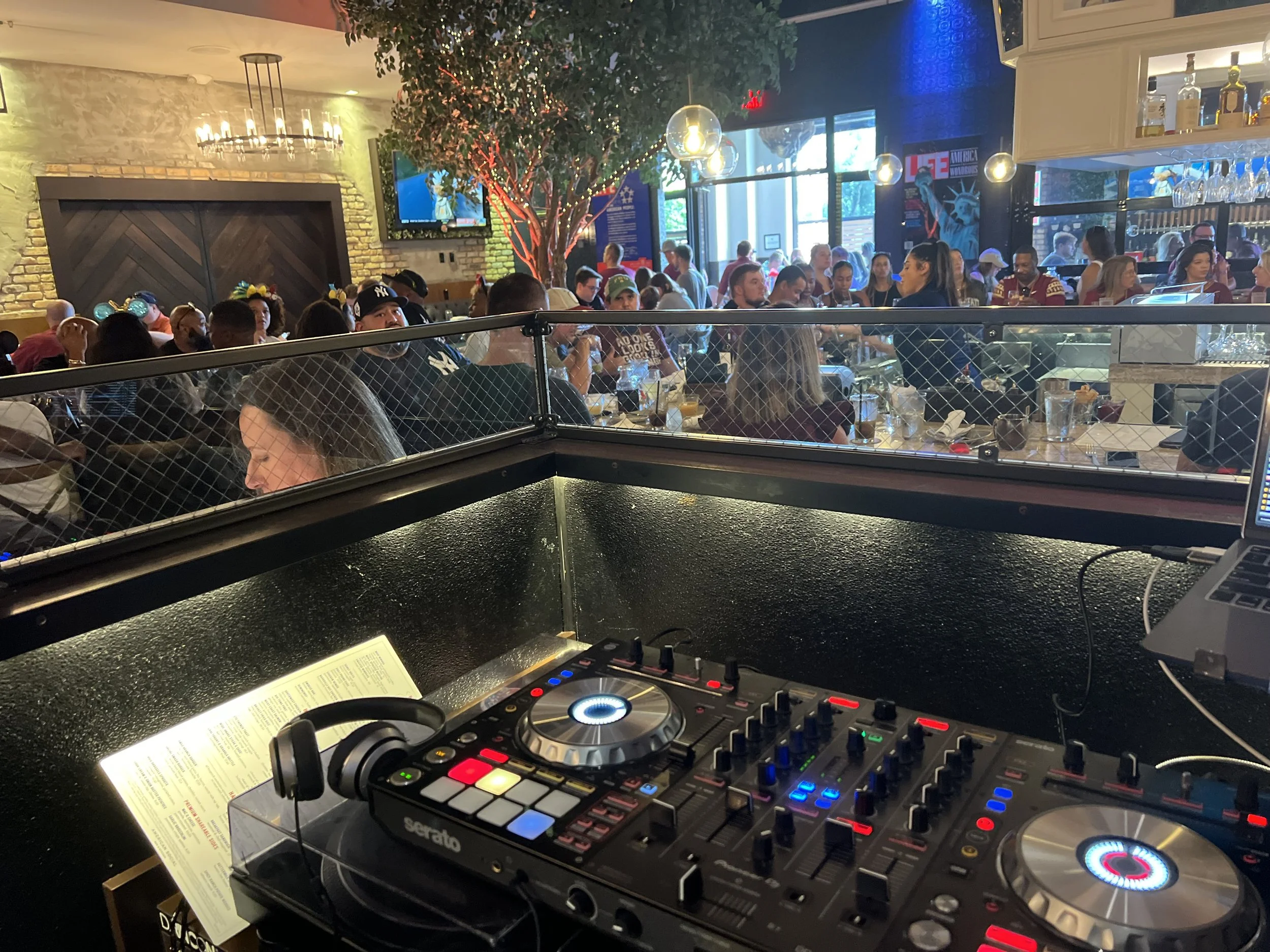 A DJ setup with turntables, a mixer, and headphones in the foreground, overlooking a crowded restaurant or bar with people dining and socializing, some wearing Mickey Mouse ears, in a lively indoor setting with decorative lighting and large windows.