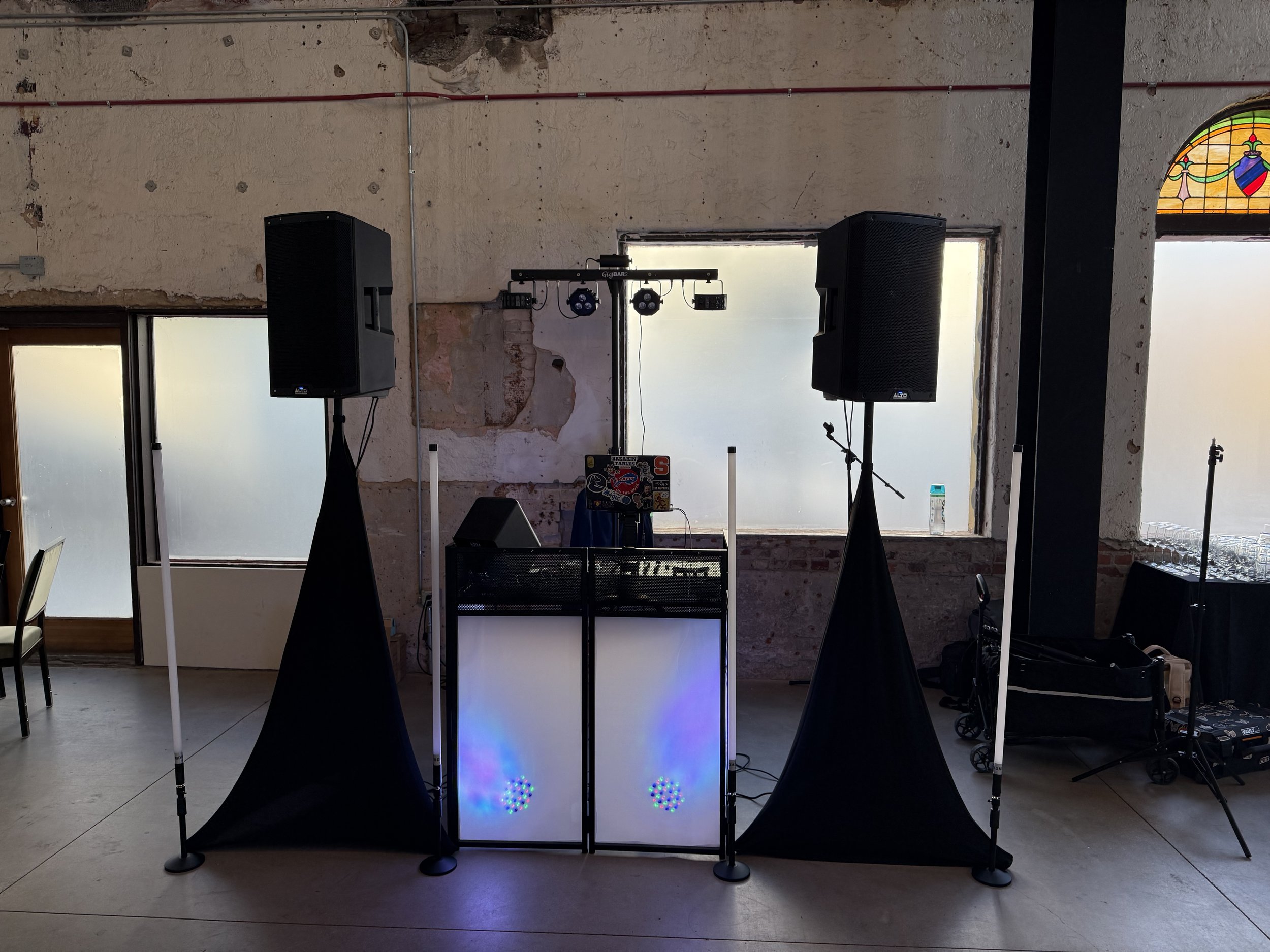 DJ booth setup with two tall speakers on stands, a DJ table with colorful lights, and fog machines on either side inside an industrial-style venue with exposed brick walls and large windows.