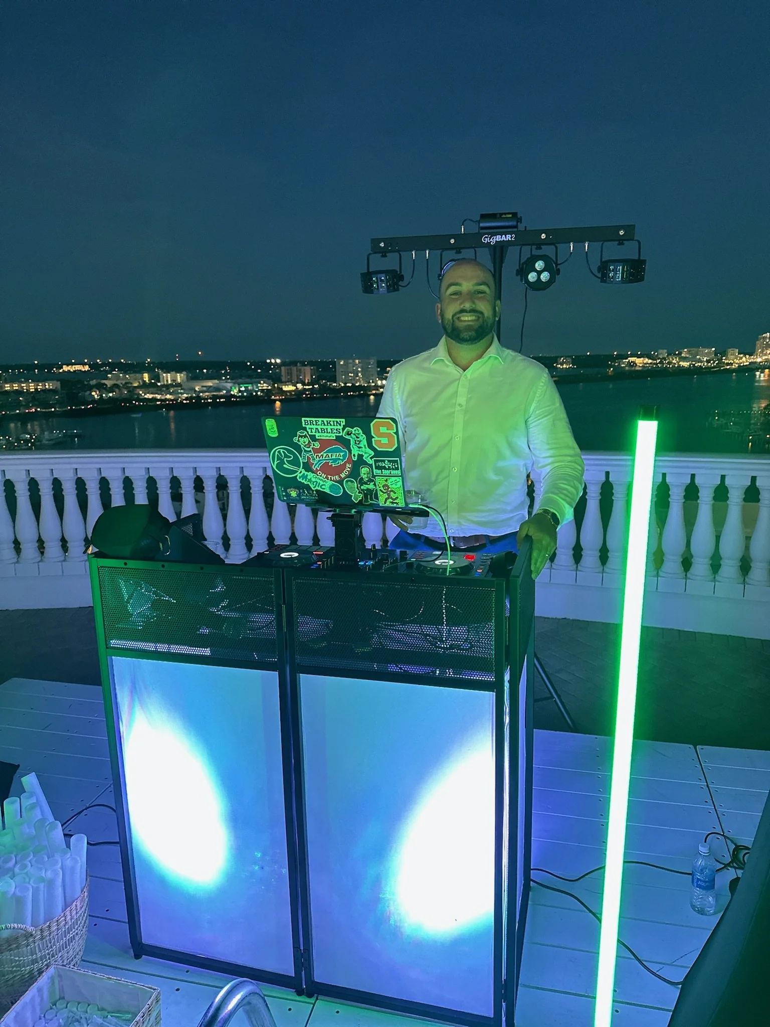 A DJ standing behind a DJ booth on a rooftop at night, with city lights and water in the background, illuminated by green and blue lights.