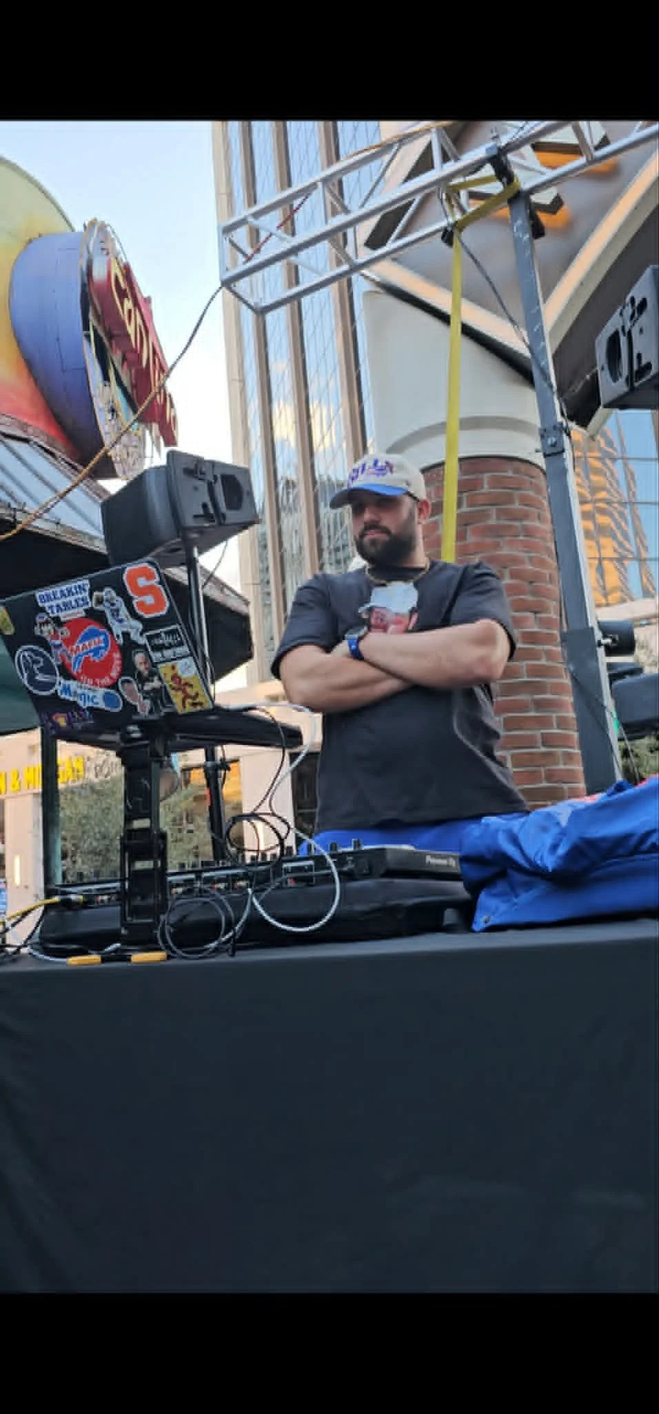 A DJ standing at an outdoor event, surrounded by sound equipment, with a city building in the background, crossed by black bars at the top and bottom.