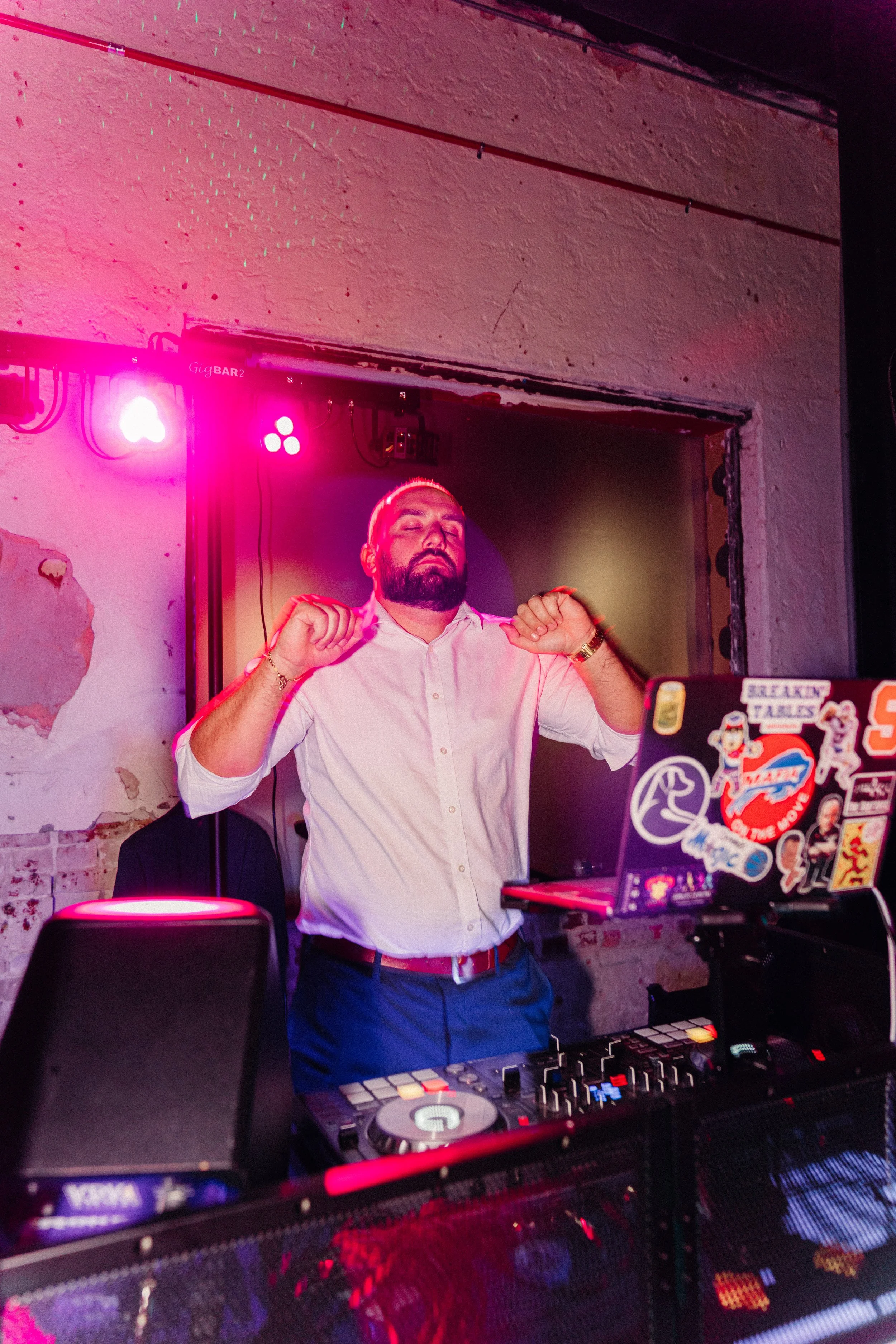 A man with a beard and closed eyes is dancing behind DJ equipment with stickers, wearing a white shirt and gold watch, in a dimly lit venue with pink and purple lights.