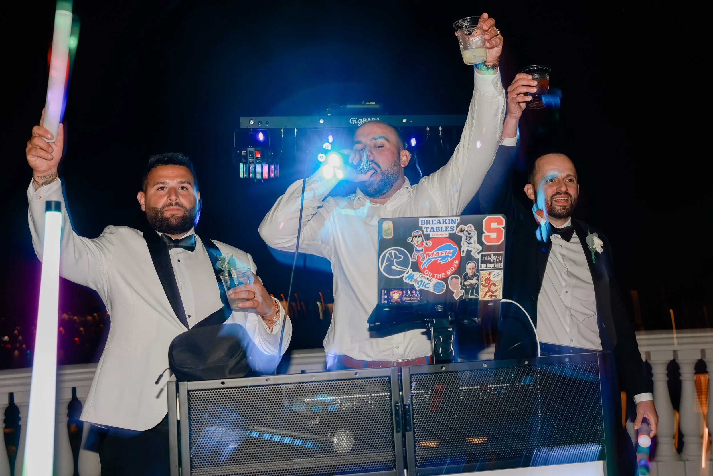 Three men in tuxedos and white shirts celebrating at a party, one singing into a microphone and the others holding drinks, in a festive setting with colorful lights.