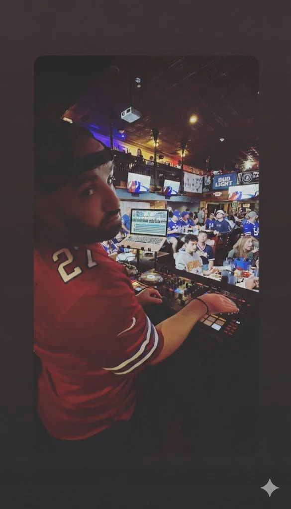 A DJ or music producer working with a laptop and mixing console in a crowded bar or sports bar with people watching a game on multiple screens.