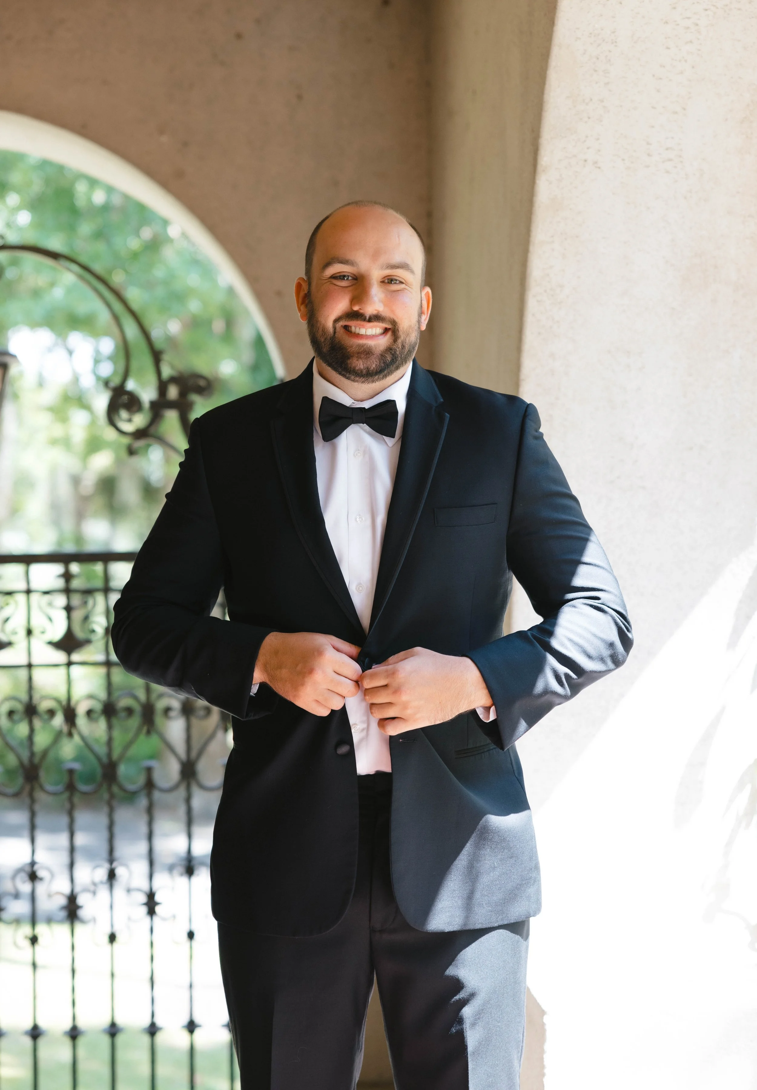 A man in a black tuxedo with a bow tie, smiling, standing on a balcony with a wrought-iron gate and greenery in the background.