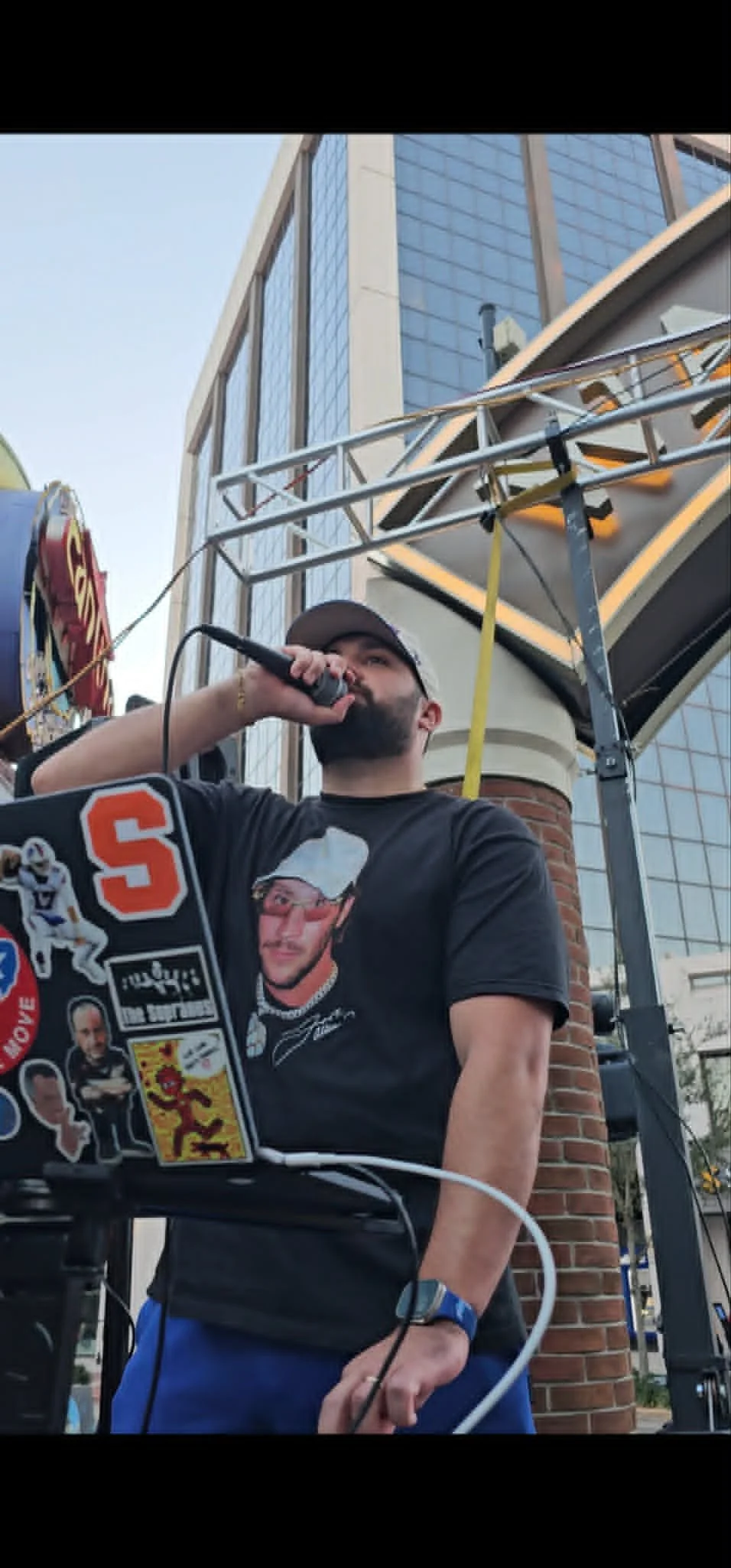A man wearing a black T-shirt and a cap speaking into a microphone outdoors, with a laptop decorated with stickers, a stage structure, and a large modern building in the background.