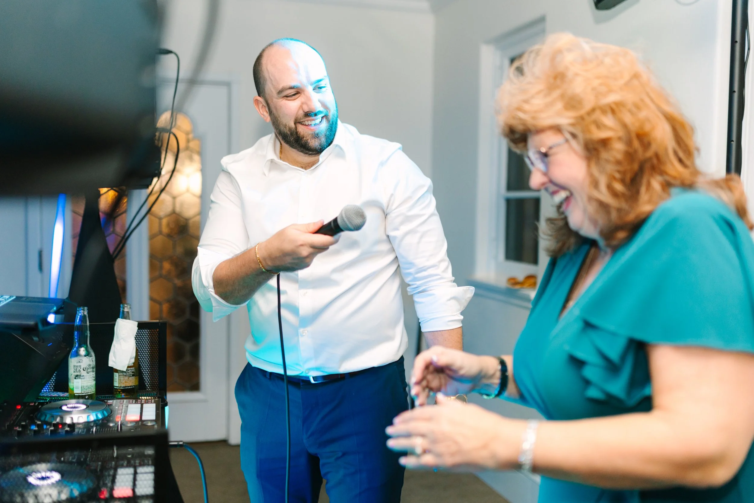 A man in a white shirt holding a microphone, smiling, and a woman in a teal dress laughing, in a room with DJ equipment and bottles on a table.