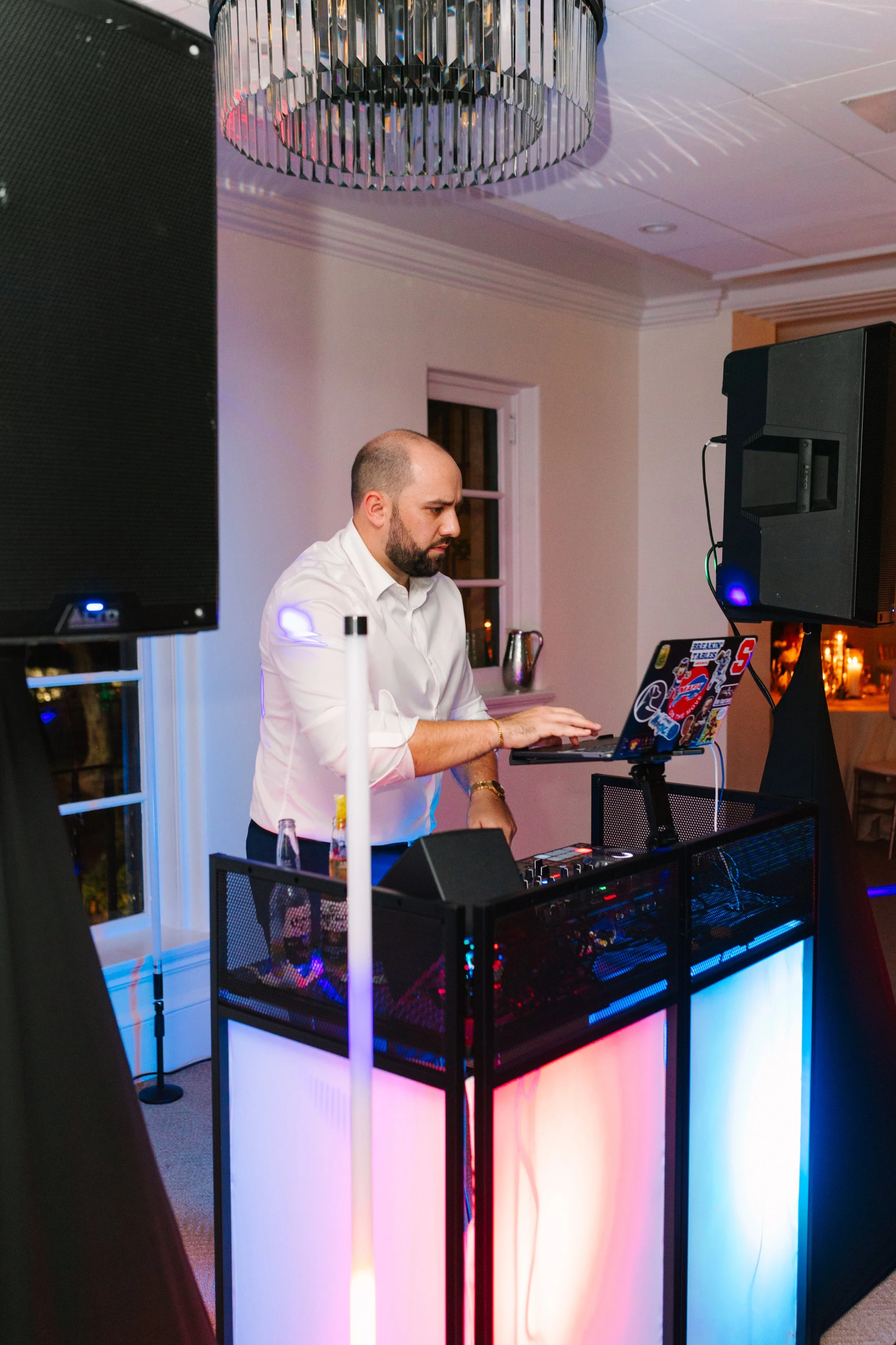 A man with a beard and wearing a white shirt DJing at an event with colorful lighting, standing behind a DJ setup with speakers and equipment in a decorated indoor venue.