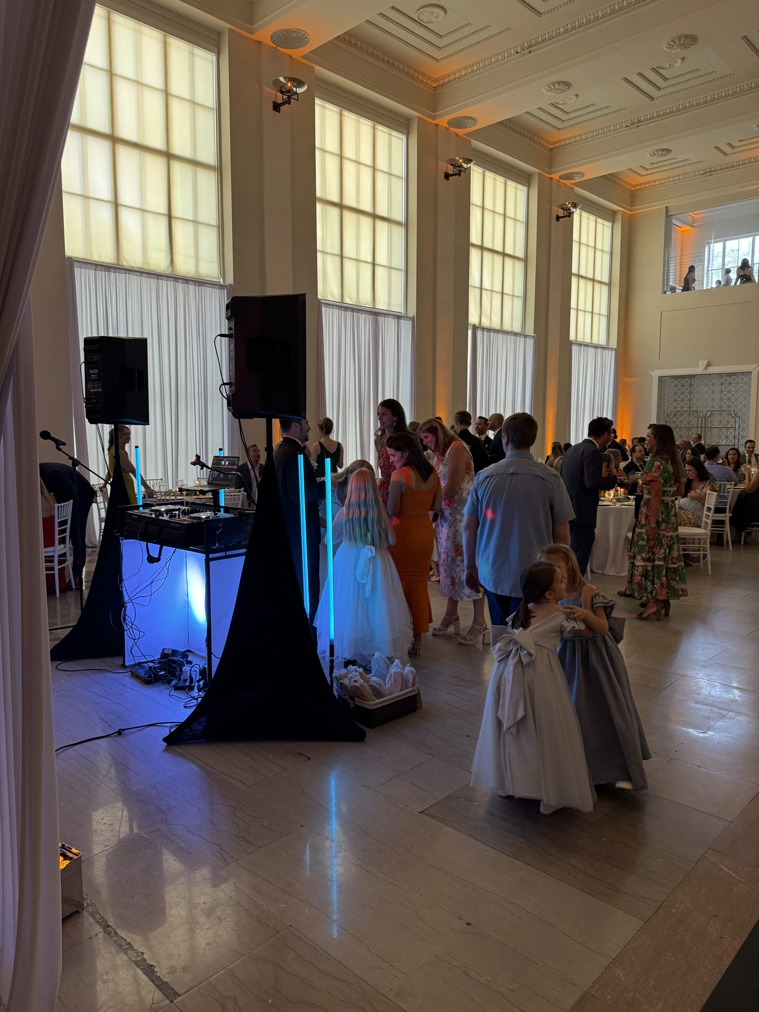 People dancing and socializing at a formal event or wedding reception in a large, elegant hall with tall windows and warm lighting.