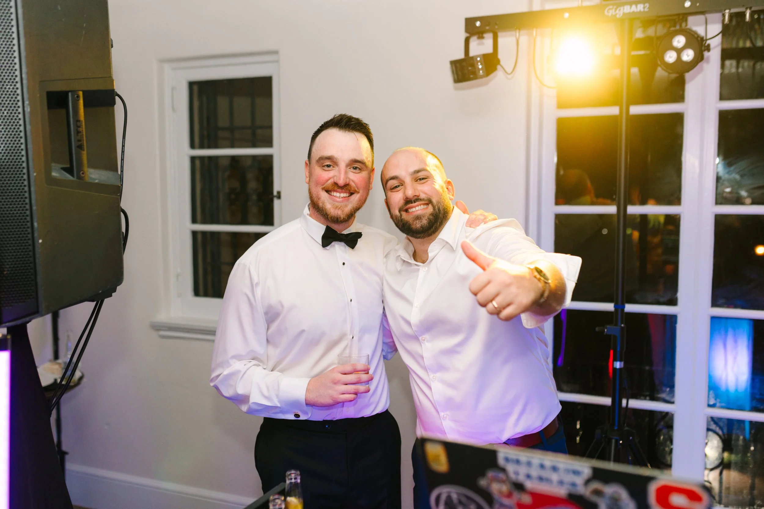 Two men dressed in formal attire, one in a tuxedo and the other in a white shirt, smiling and posing for a photo indoors at a social event or party, with lights and equipment in the background.