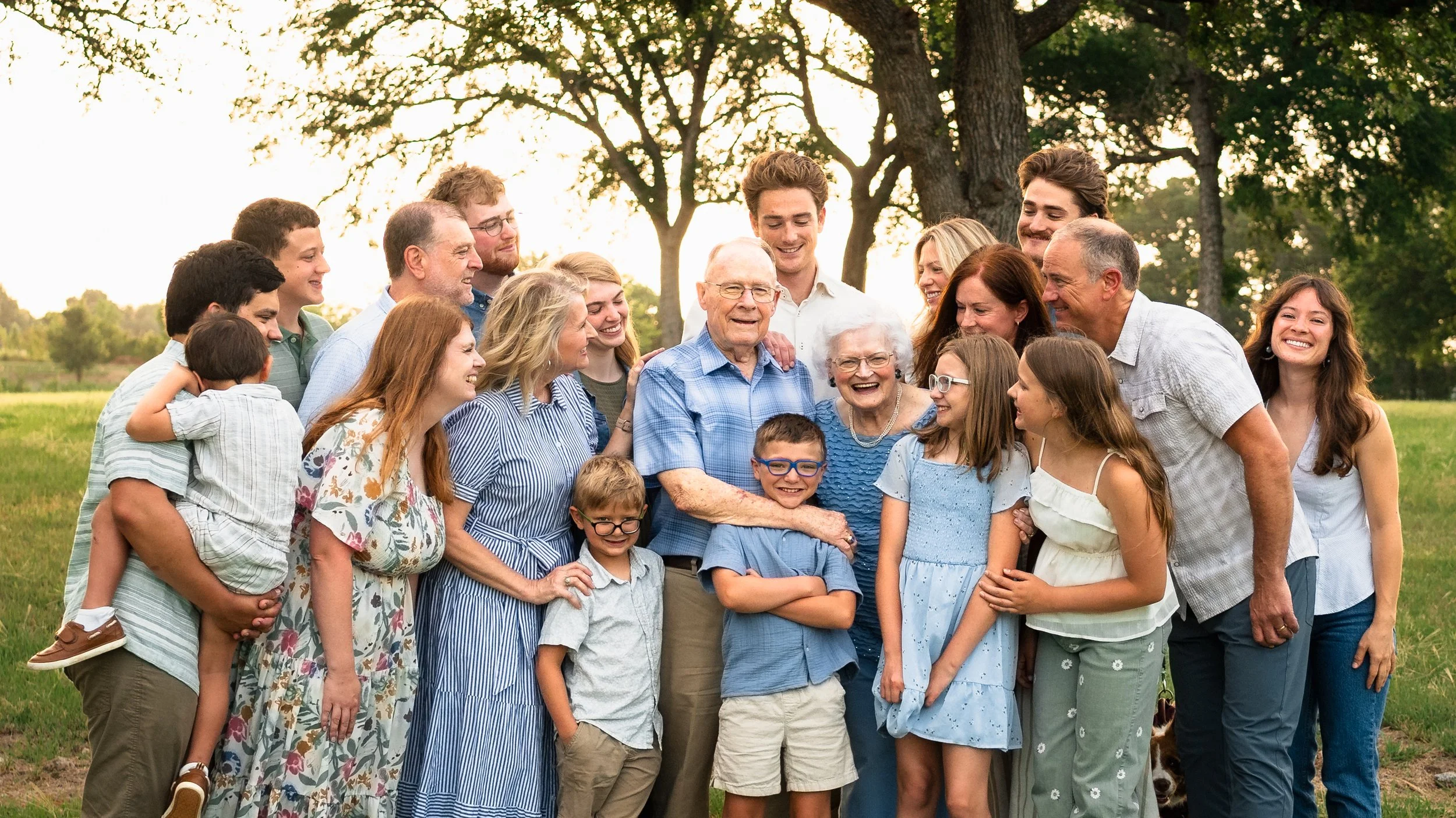 A large multigenerational family group photo outdoors in a park with trees in the background, showing smiling faces of children, adults, and elderly individuals gathered together.