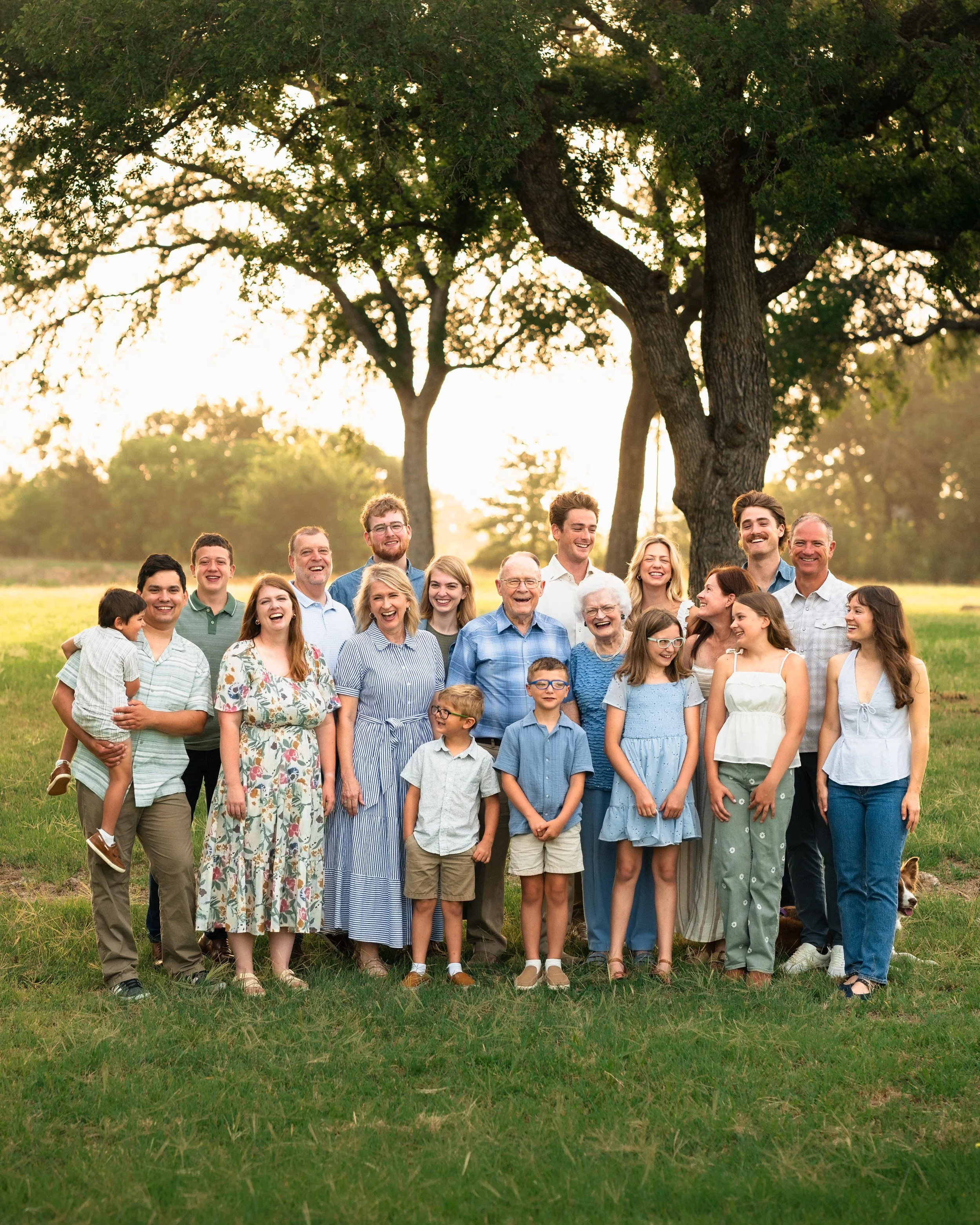 A large group of diverse people of various ages standing outdoors on grass, smiling, with trees and the sun setting behind them.